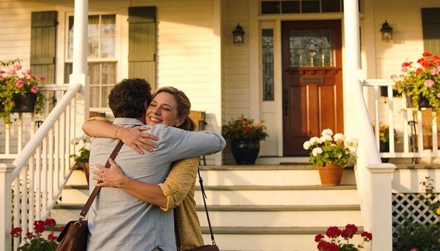 Couple embracing on sunlit front porch with blooming pots, warm homecoming moment