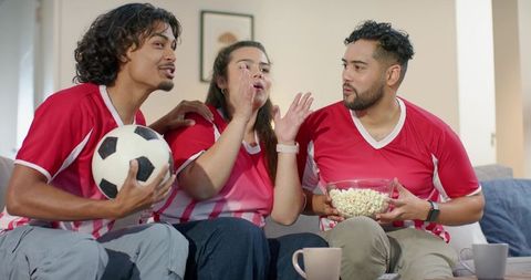 Three friends cheering during soccer match on sofa wearing red jerseys and eating popcorn