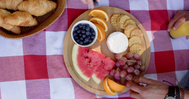 Beautiful picnic setup with a variety of fruits including watermelon, blueberries, grapes, and orange slices. Also shown are crackers with a cheese wheel and croissants. All arranged on a round wooden platter on a red checkered tablecloth. This image is ideal for promoting outdoor dining ideas, picnic events, food blogs, and summer lifestyle advertisements.