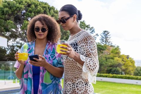African american friends poolside sharing smartphone and citrus drinks in stylish coverups