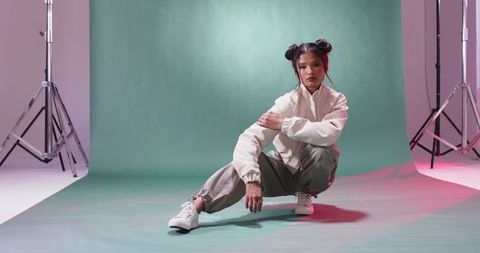 Crouching asian woman in streetwear with bomber jacket and sneakers on green backdrop