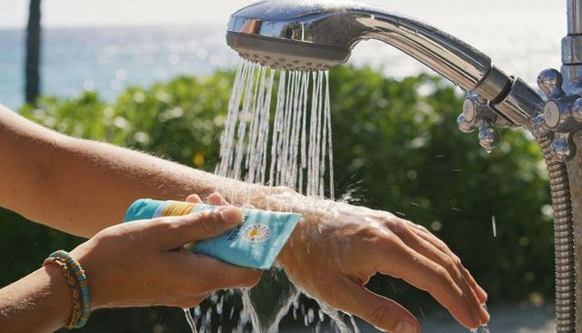 Applying sunscreen and rinsing hands under chrome beach shower with ocean background and water spray