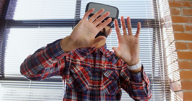 A man is interactively engaging with a virtual reality headset in a bright indoor environment, wearing a plaid shirt. He is extending his hands forward, possibly interacting with virtual objects. This visual can be used to illustrate concepts in technology, augmented and virtual reality developments, user experiences in VR environments, or future technology advancements.