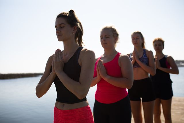 Group of four Caucasian women in sportswear standing in a row on a jetty by the river, practicing yoga and meditation with eyes closed. Ideal for use in articles or advertisements related to fitness, team building, mindfulness, outdoor activities, and wellness retreats.