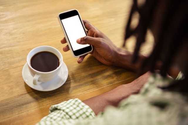 Person holding smartphone with blank screen while sitting at wooden table with cup of coffee. Ideal for illustrating themes of technology, relaxation, casual lifestyle, and communication. Suitable for use in blogs, social media posts, advertisements, and articles about modern lifestyle, coffee culture, and mobile technology.