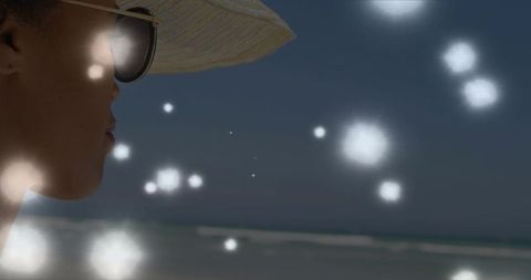 Gazing woman wearing straw hat and sunglasses at twilight beach with dreamy bokeh lights