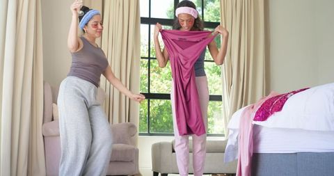 Friends trying on magenta dress while wearing skincare patches and headbands in bedroom
