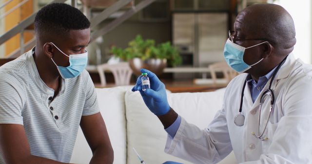 African american senior male doctor giving covid vaccine to male patient in home, wearing face masks. healthcare at home in isolation during coronavirus covid 19 pandemic.