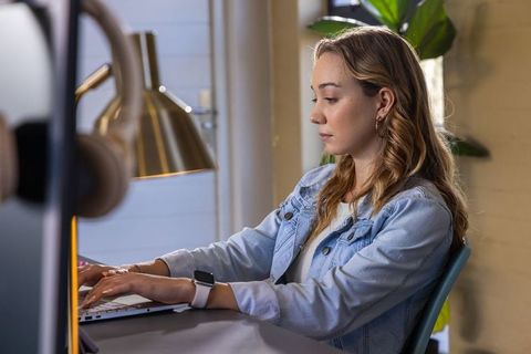 Young woman typing on laptop at home desk wearing denim jacket and smartwatch