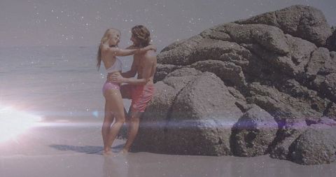 Young couple embracing on sunlit beach by rocky outcrop with lens flare and reflection