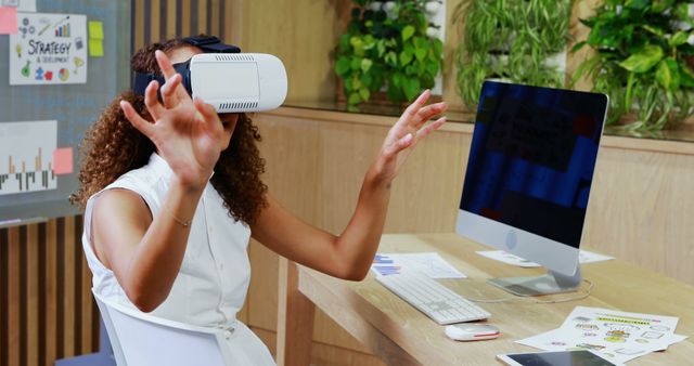 Individual using virtual reality headset at desk in a modern office environment. Can be used for themes related to advanced technology in workspaces, innovative office setups, digital work trends, and the impact of VR in professional settings.