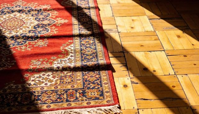 Sunlit oriental rug on worn parquet floor casting shadows, curled edge, medallion detail