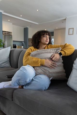 Contemplative african american woman on sofa hugging pillow checking smartwatch