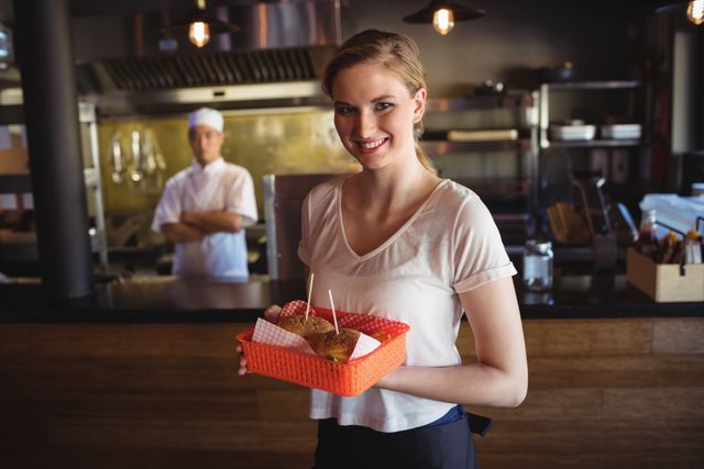 Smiling waitress holding a tray with a burger in a restaurant, with a chef in the background. Ideal for use in advertisements for restaurants, food service training materials, hospitality industry promotions, and customer service brochures.