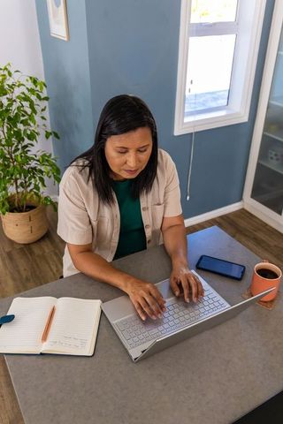 Mature woman working from home typing on laptop at desk with notebook phone and coffee