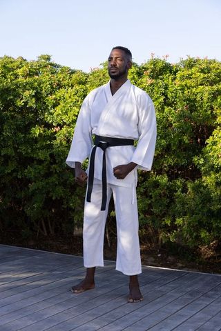 An African American man stands poised in a martial arts stance on a wooden deck surrounded by lush greenery. Dressed in a traditional white gi and black belt, he exemplifies strength, discipline, and focus. This image captures the essence of outdoor fitness and well-being, making it ideal for use in materials promoting health, exercise routines, or martial arts courses. Perfect for illustrating on topics related to wellness, nature-based exercises, or showcasing diversity in sports.