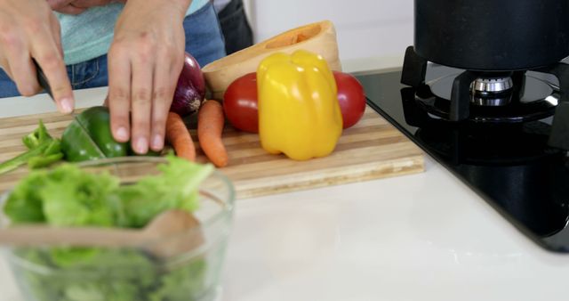 Person cutting fresh vegetables on wooden cutting board in modern kitchen. Vibrant bell peppers, carrots, and leafy greens add color to the scene. Ideal for recipes, meal preparation blogs, cooking tutorials, and promoting healthy eating.