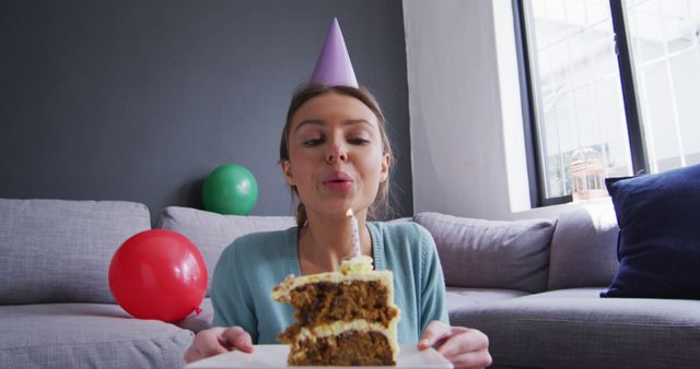 An image showing a woman in a light blue sweater and a party hat blowing out a candle on a birthday cake while sitting in a cozy living room. The background features a gray wall, a sofa, and colorful balloons, suggesting a festive atmosphere. This image can be used for birthday cards, party invitations, or advertisements for birthday-related products and celebrations. It evokes feelings of joy and the warmth of home celebrations.