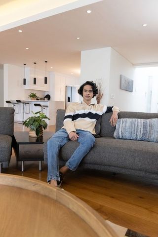 Asian man sitting on gray sofa in modern open-plan living room with kitchen island