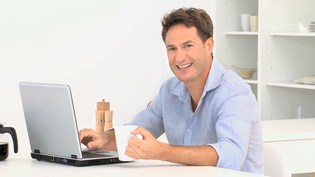 This image shows a man sitting in a bright kitchen holding a coffee mug and smiling towards the camera while working on his laptop. This can be used for articles or websites focusing on remote work, home office setups, relaxed working environments, or lifestyle blogs emphasizing work-life balance.