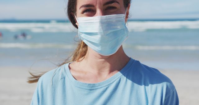 Woman wearing a face mask standing on a sunny beach. Background shows ocean waves and a clear sky. Useful for health and safety promotions, outdoor activities during pandemic, travel safety guidelines, and beach outing ads.