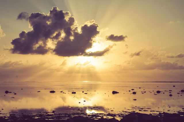This image features a golden sunrise with a striking cloud formation over calm ocean waters. The sunrays filter through the clouds, creating a beautiful reflection on the water's surface. Ideal for use in motivational posters, travel brochures, nature-themed websites, or as a calming background in digital content.