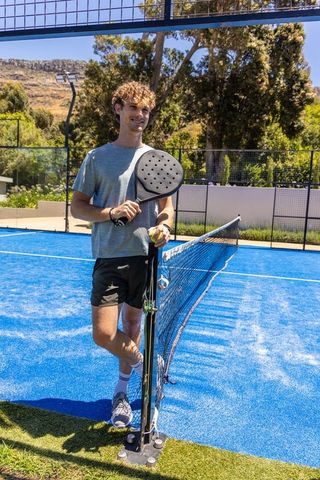 Young man resting against net post gripping padel racket and ball on bright blue court