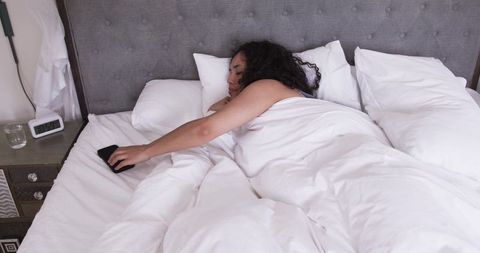 African american woman reaching for smartphone while sleeping in cozy white bed