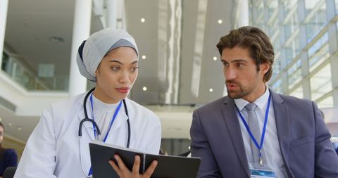 image of female doctor in a white coat discussing ideas with a businessman at a conference setting. they are using a digital tablet to go over details, indicating collaboration between medical and business professionals. captures the importance of cross-industry partnerships and information sharing. suitable for articles, presentations or promotional material highlighting corporate events, forums on innovation, or expert consultations.