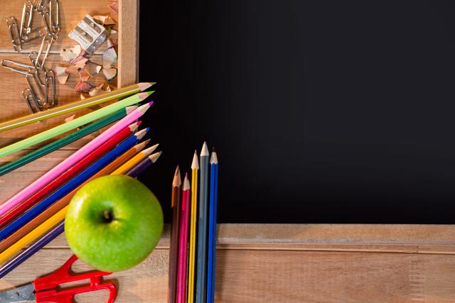 Colorful pencils and a green apple placed on a wooden desk next to a chalkboard. Ideal for educational materials, back-to-school promotions, classroom decor, and creative projects. Perfect for illustrating concepts related to learning, teaching, and school environments.