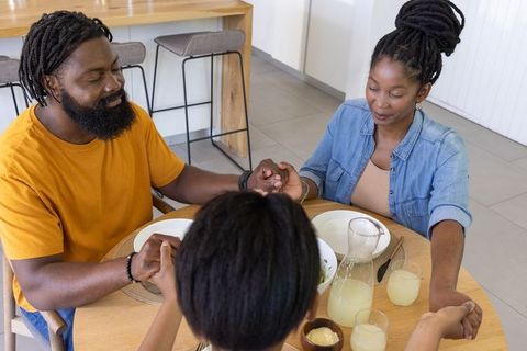 African american family holding hands and praying together before meal at kitchen table