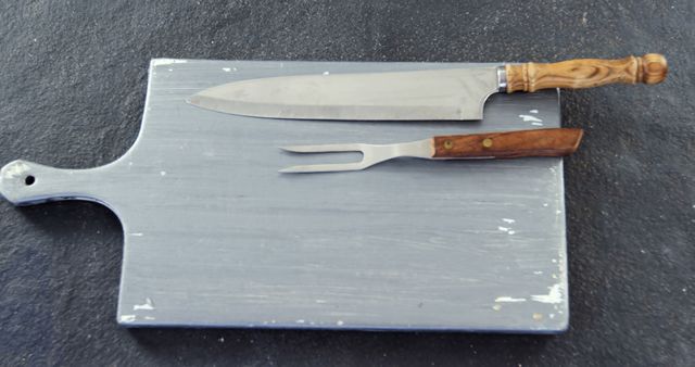 A chef's knife and a carving fork rest on a worn cutting board, with copy space. These tools are essential for food preparation and presentation in culinary settings.