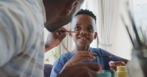 African american father painting smiling son during home craft time with bright paints