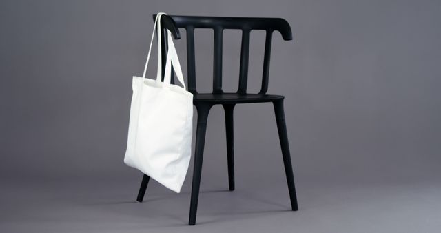 A white tote bag hangs on a sleek black chair against a neutral background, with copy space. Its simplicity suggests a minimalist aesthetic or could symbolize sustainable fashion choices.