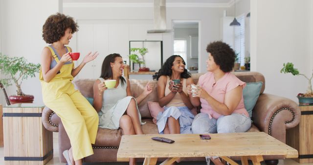 Four women sitting on a comfortable sofa in a living room, smiling and chatting while holding cups of coffee. They are in a casual and relaxed atmosphere, enjoying each other's company. The scene includes a wooden coffee table with a TV remote on it and potted plants, enhancing the cozy and inviting look. Ideal for use in lifestyle, friends gathering, coffee culture, and home decor contexts.