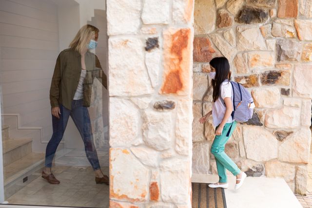 Caucasian mother opening the door as her daughter is walking up the front door while wearing a backpack. both of them are wearing masks.
