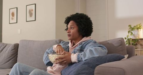African-american man watching movie on sofa eating popcorn in cozy denim jacket
