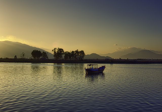 Lonely fishing boat floating on calm lake water during sunset with silhouettes of trees and mountains in the background. Sky glows with soft shades of blue and yellow from setting sun. Ideal for promoting travel destinations, nature conservation, outdoor activities, peaceful retreats.