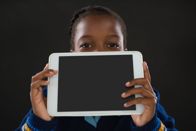 Young schoolgirl holding a digital tablet in front of her face against a black background. This image can be used for educational content, technology in education, online learning, or advertisements for digital devices. It is suitable for illustrating concepts related to modern education, children's use of technology, and playful learning environments.