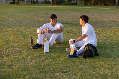 Two cricket teammates sitting and chatting on field during golden hour practice break