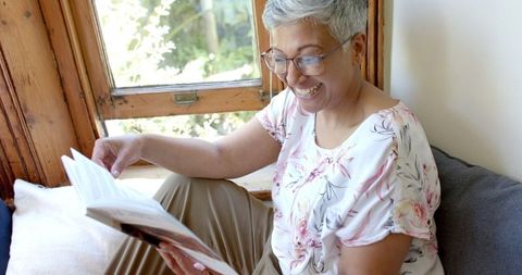 Happy Senior Woman Reading Book by Sunny Window at Home
