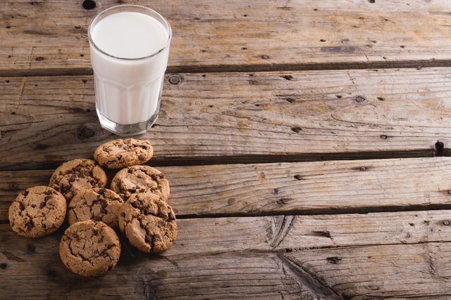 High angle view of chocolate chip cookies next to a glass of milk on a rustic wooden table. Perfect for illustrating concepts of comfort food, homemade snacks, or healthy eating. Ideal for use in food blogs, recipe websites, or advertisements for dairy products and baked goods.