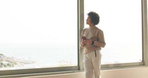 African american man standing by ocean-view loft window holding smartphone, contemplative