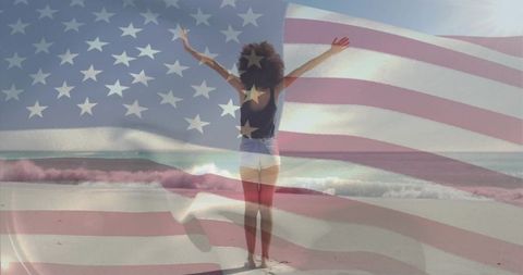 Woman raising arms on sunlit beach with translucent american flag overlay, patriotic freedom concept