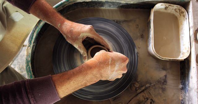 Hands are shaping wet clay on a potter's wheel. The image focuses on the delicate and skilled action involving the artisan's hands as they craft a pottery piece. Ideal for promoting artistic and handcrafted goods, illustrating pottery-making processes, or emphasizing craftsmanship and creativity in branding materials.