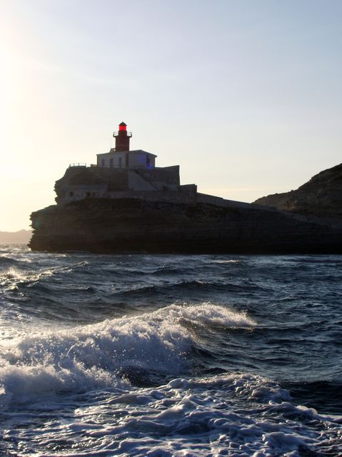 Lighthouse perched on a rocky coastal cliff during sunset with ocean waves crashing below. Ideal for travel brochures, maritime navigation guides, and coastal photography. Evokes a sense of solitude, navigation, and the power of nature.