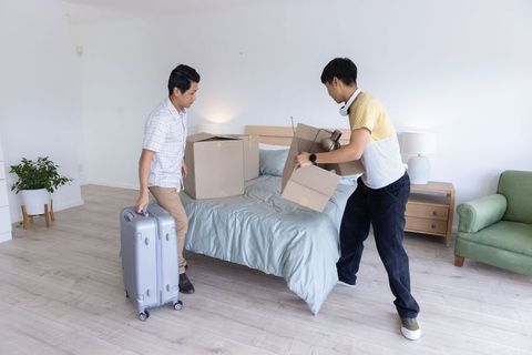 Asian men moving into apartment carrying boxes and suitcase in bright minimalist bedroom