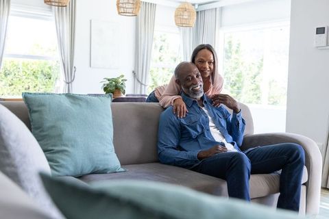 Senior couple relaxing on sunlit living room sofa, affectionate mature partners smiling