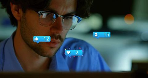 Young man concentrating on laptop late night with social media overlays and glasses
