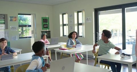 Diverse teenagers collaborating in modern classroom using laptops, tablets, notebooks